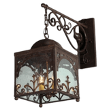 A wrought iron sconce features intricate scrollwork and a square glass enclosure, casting a warm glow from inner candle-like bulbs.