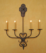 A decorative wrought iron wall sconce featuring intricate curves and elegant candle holders. The design highlights a classic style with four slender arms extending from a central point, topped with lighted candles, set against a warm, textured background.