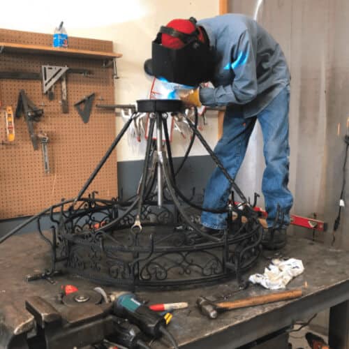 A skilled artisan welds a custom wrought iron chandelier in a workshop. The intricate design features ornate scrollwork and a sturdy base, showcasing craftsmanship in the lighting fixture. Tools and equipment are visible in the background, highlighting the production process.