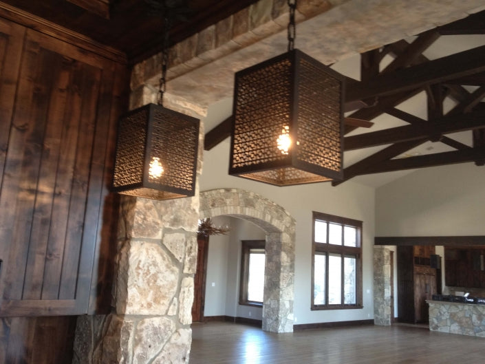 Two intricately designed wrought iron pendants hang from a wooden ceiling, illuminating a spacious room with stone accents and large windows. The warm glow enhances the rustic charm of the interior.