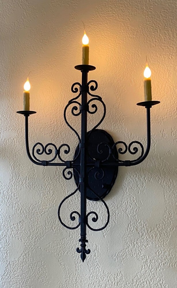 A black wrought iron wall sconce featuring intricate scroll designs and three candle holders. The warm glow of candle-like bulbs creates a welcoming ambiance, enhancing the decorative elegance of any space.