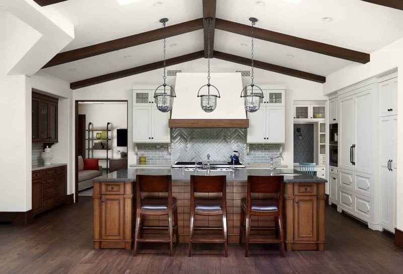 Three elegant wrought iron pendant lights hang above a modern kitchen island with a dark wood finish. The space features light cabinets, white tiles, and exposed wooden beams, creating a warm and inviting atmosphere. The fixtures add a rustic charm, complementing the stylish cabinetry and countertops.
