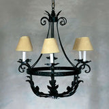 A decorative chandelier featuring a circular wrought iron frame adorned with intricate leaf designs. It houses three elegant cream-colored lampshades that gently diffuse light, adding warmth to any space. The design combines elegance and artistry, perfect for enhancing traditional or rustic interiors.