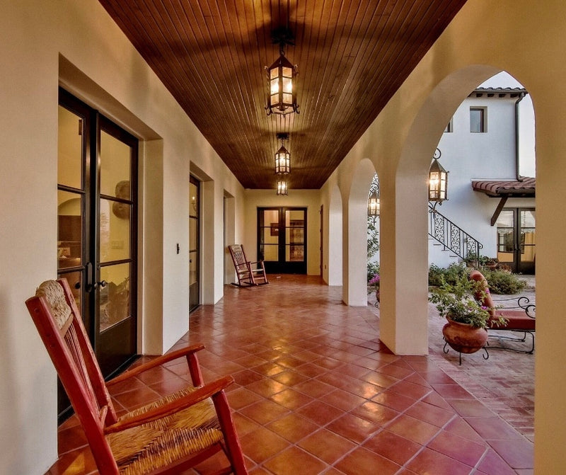 A cozy outdoor corridor featuring elegant wrought iron lighting fixtures hanging from a wooden ceiling. The rustic design complements the terracotta tiled floor and soft archways, creating an inviting atmosphere. Two wooden rocking chairs sit on one side, enhancing the warm, welcoming feel of the space.